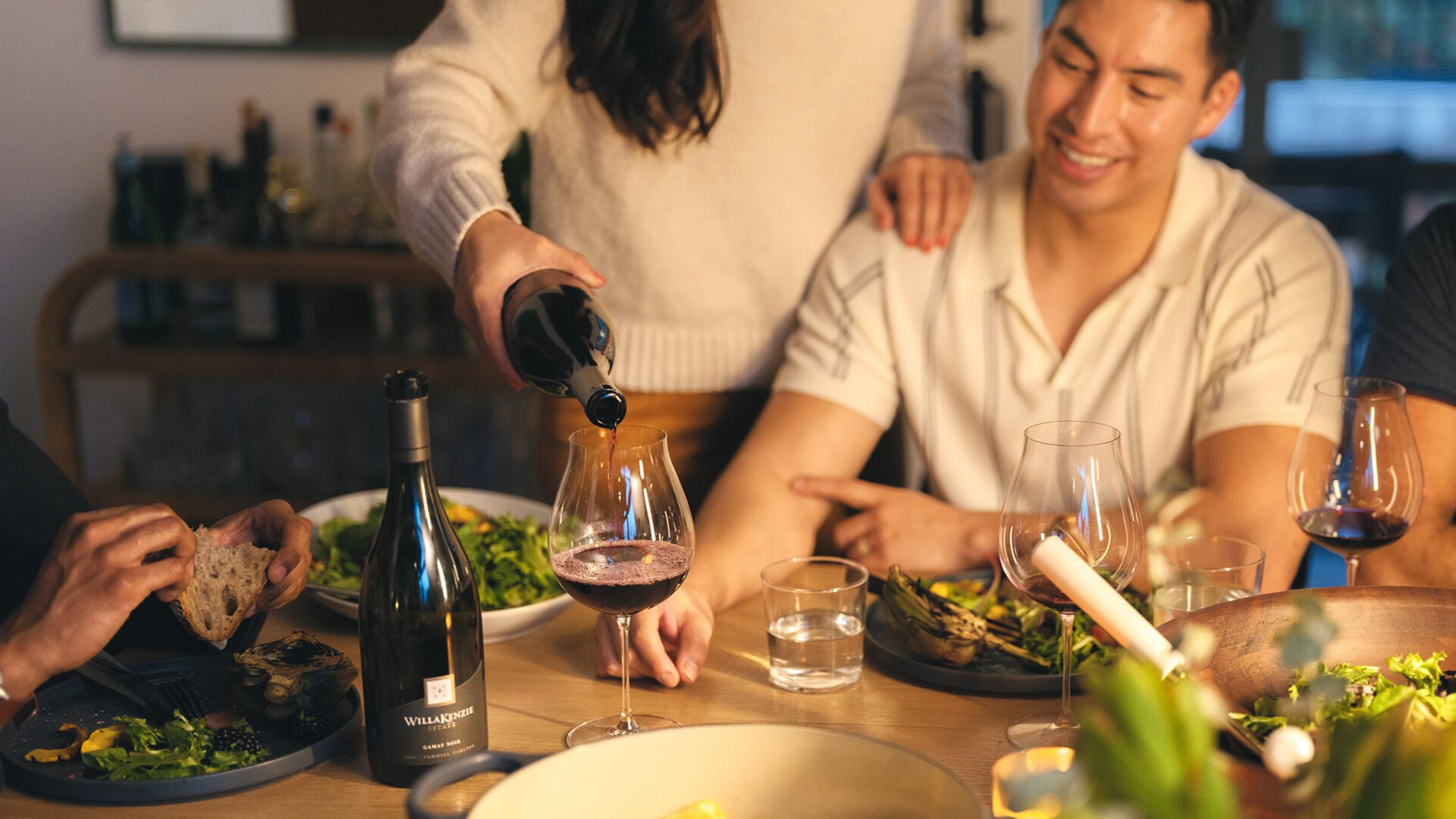 Dinner party with woman pouring gamay noir over man's shoulder in glass.